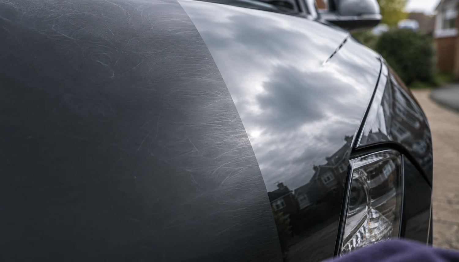 Close-up of a dark car's front wing split down the middle: dull, swirl-marked paint on one side and a machine-polished mirror finish on the other, reflecting the sky and houses behind